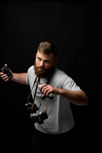 Fotógrafo barbudo bonito em uma camiseta cinza com um monte de câmeras diferentes em uma mão e em um ombro olhando para uma câmera e pronto para fazer um bom tiro. — Fotografia de Stock