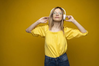 Sarı tişörtlü, beyaz kulaklıklı, müzik dinleyen, dans eden ve mavi arka planda gülen güzel sarışın kadın. Rahatlamak ve eğlenmek. Yaşam biçimi.