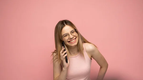 Foto close-up de atraente positiva rindo jovem loira vestindo camisa amarela elegante e óculos se comunicando no telefone celular e sorrindo isolado sobre fundo amarelo. — Fotografia de Stock