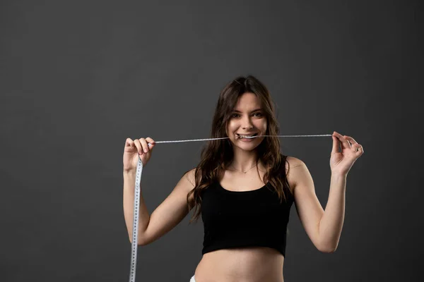 Ung brunett fitness kvinna i en svart topp med en måttband i munnen. Hälsovård och hälsosam kost. Perfekt smal kropp. — Stockfoto
