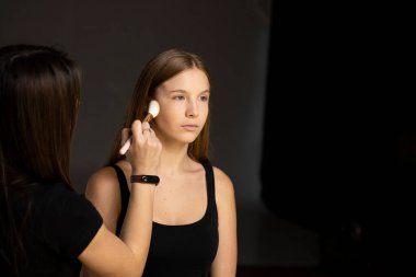 Model yüzünde fırçayla çalışan profesyonel bir makyöz. Makyaj yapma süreci. Güzellik salonunun içinde sarışın bir kadının portresi. Cilde ton uygulanıyor.