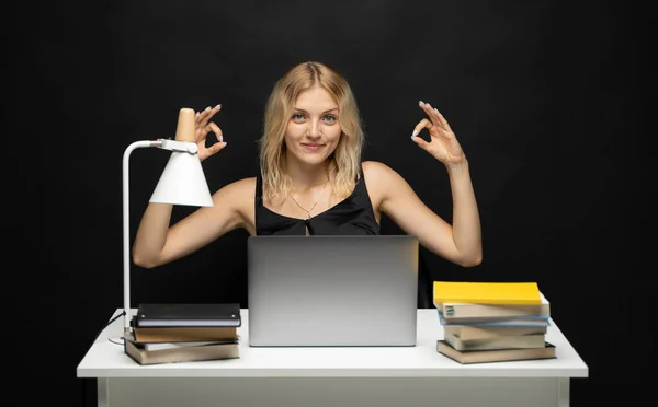 Mujer rubia alegre en camisa negra que trabaja con un ordenador portátil y libros y muestra gesto signo ok a un cliente o colegios sobre una charla. Oficina de trabajo. — Foto de Stock