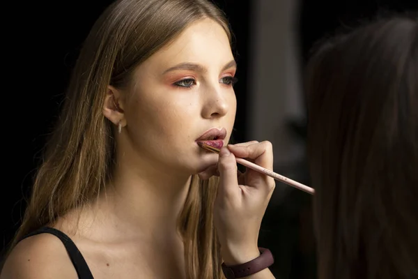 Make-up malíř nanáší červenou rtěnku na ženskou modelku. Ruka maskérky, malování rtů mladé modelky krásy. Doplnit v procesu. — Stock fotografie