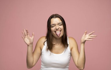 Beyaz tişörtlü esmer bir kadının parlak çılgın portresi pembe arka planda gözleri kapalı bir dille bağırıyor. Neşeli komik stil, sportif bronzlaşmış vücut. Duygular.