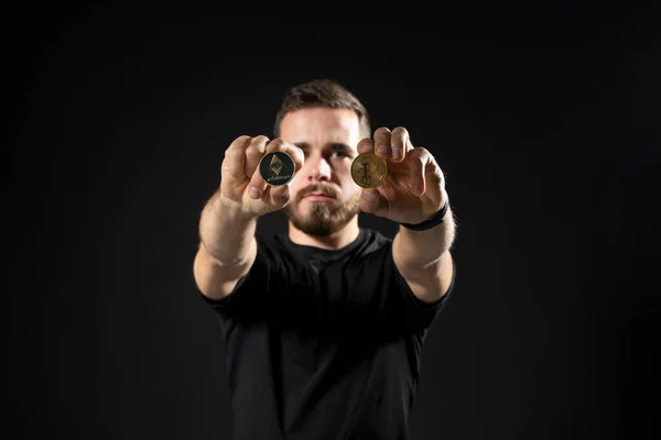 Молодий бородатий чоловік, що тримає віртуальну валюту ethereum монету і біткоїн на чорному тлі студії. Криптовалюта. — стокове фото