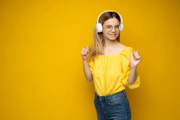 Hermosa mujer rubia joven atractiva con camiseta amarilla y gafas en auriculares blancos escuchando música, bailando y riendo sobre fondo amarillo en el estudio. Estilo de vida. —  Fotos de Stock