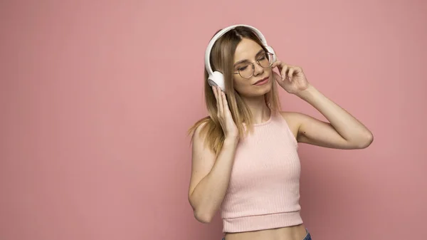Linda chica con una camiseta rosa y gafas escuchando música con sus auriculares y bailando sobre fondo rosa. —  Fotos de Stock