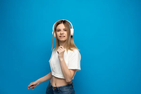 Jovencita alegre bonita sonriendo mientras escucha música en auriculares y bailando sobre fondo azul. —  Fotos de Stock