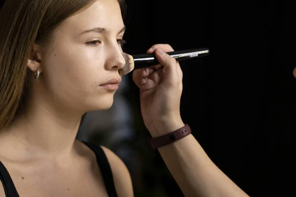 Processen med at gøre makeup. Make-up artist arbejder med pensel på model ansigt. Portræt af ung blond kvinde i skønhedssaloon interiør. Anvendelse tone på huden . - Stock-foto