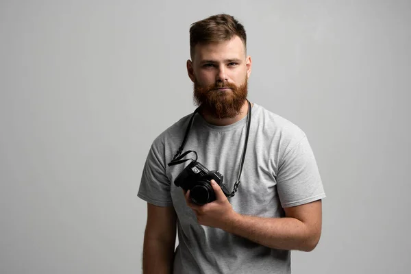 Retrato de fotógrafo profesional barbudo con cámara dslr mira directamente a la cámara aislada sobre fondo gris. —  Fotos de Stock