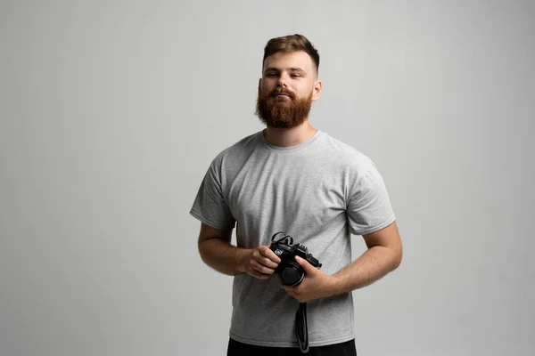 Portret brodatego profesjonalnego fotografa w szarej koszulce z kamerą dslr patrzy prosto w kamerę odizolowaną na szarym tle. — Zdjęcie stockowe