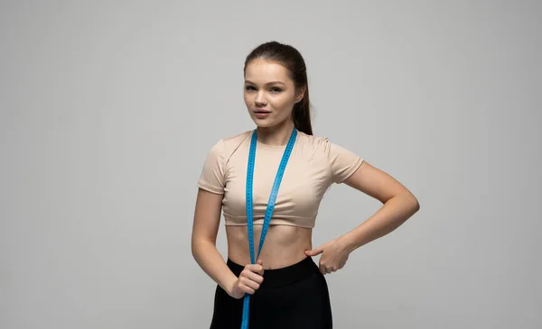 Schöne schlanke, sportliche Frau in Sportbekleidung mit blauem Maßband um den Hals und blickt geradeaus in eine Kamera auf weißem Hintergrund. Sport und Ernährung. — Stockfoto