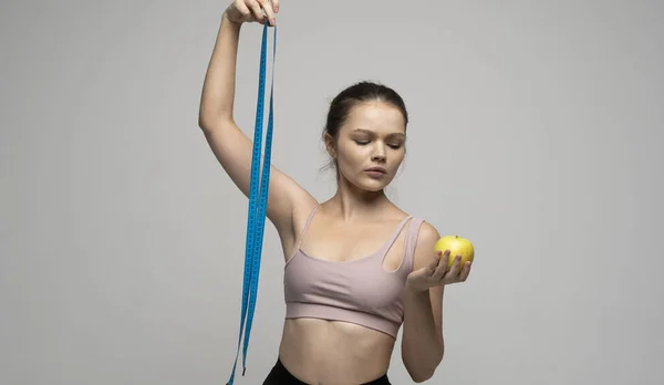 測定テープで立っているスポーツ衣装の魅力的なセクシーなブルネットの女性を混乱させ、白い背景にリンゴを保持しています。美しさと健康の概念。健康的な食事と食事. — ストック写真