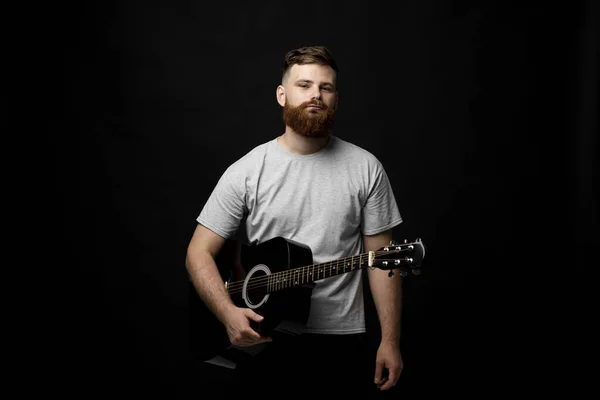 Красива брюнетка бородатий музикант стоїть і тримає акустичну гітару в руці і дивиться в камеру на чорному фоні студії. Готовий відтворювати музику . — стокове фото