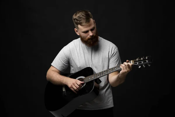 Гарна брюнетка бородатий гітарист грає на акустичній гітарі в чорній кімнаті. Концепція запису музики, репетиції або живого виконання . — стокове фото