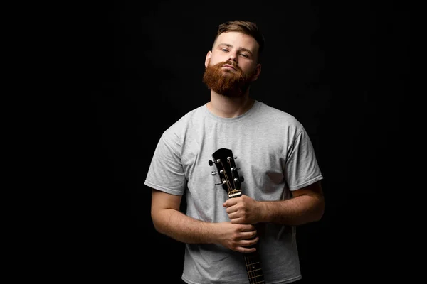Портрет красивої брюнетки бородатий музикант, гітарист стоїть і тримає акустичну гітару в руці і дивиться в камеру на чорному фоні студії. Готовий відтворювати музику . — стокове фото