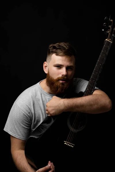 Портрет красивої брюнетки бородатий музикант, гітарист тримає акустичну гітару в руці і дивиться в камеру на чорному фоні студії. Готовий відтворювати музику . — стокове фото