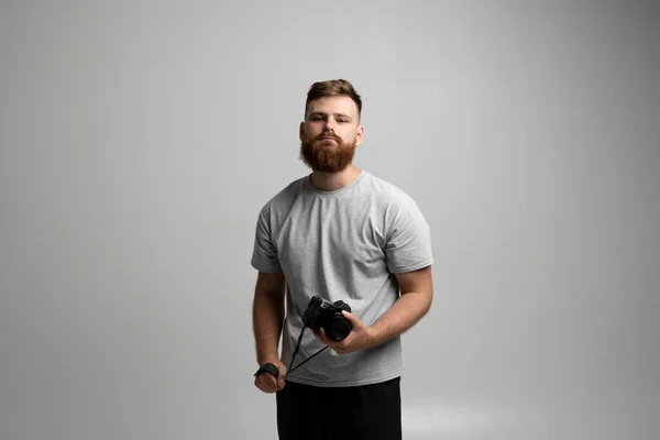 Portrait of bearded professional photographer with dslr camera looks straight into the camera isolated on gray background. — Stock Photo, Image