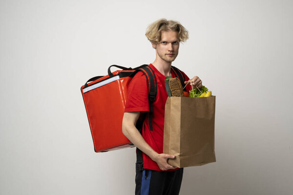 Food delivery service. Portrait of pleased delivery man in red uniform smiling while carrying paper bag with food products isolated over white background.