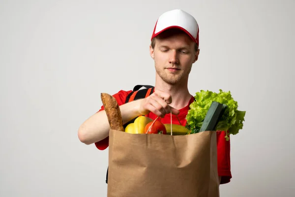 O mensageiro de entrega de supermercado homem em um gorro vermelho e t-shirt com uma bolsa térmica em uns ombros mantém o saco de papel com a comida no fundo de estúdio branco. Serviço de entrega de alimentos, compras online. — Fotografia de Stock