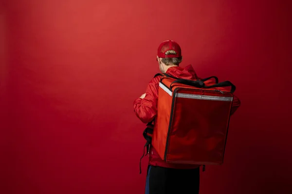 Leverans man i röd uniform med en termisk ryggsäck isolerad på en vit bakgrund. Snabb hemleverans. Beställning online. Courier levererar livsmedel hem. — Stockfoto