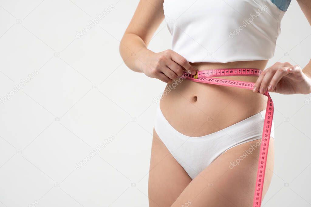 Crop image of attractive sexy sporty slim woman body in a white underwear with measuring tape around the waist. Healthy nutrition and weight losing concept. Diet concept.