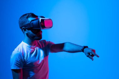 Siyah tişörtlü, 3D kulaklık takan bir adamın portresi. Sanal gerçeklikte video oyunları oynarken yaptığı bir şeye dokunur gibi el kol hareketi yapıyor. Metaevren.