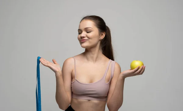 Morena mulher atraente em um desgaste esportivo é sorriso e olha em uma câmera enquanto segura fita métrica azul e uma maçã. Saúde e nutrição saudável. Corpo magro perfeito. — Fotografia de Stock