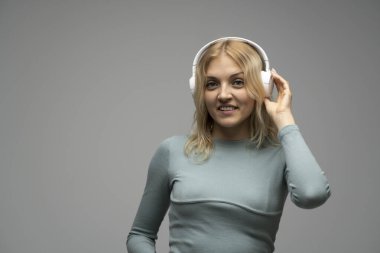 Beyaz kulaklıklı sarışın kız stüdyodaki gri arka planda kapalı gözlerle müzik dinliyor. Gri tişört giyiyor..