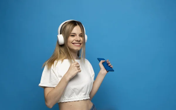 Hermosa joven rubia con auriculares y dispositivo móvil escuchando música y sonriendo y bailando, aislada sobre fondo azul. —  Fotos de Stock