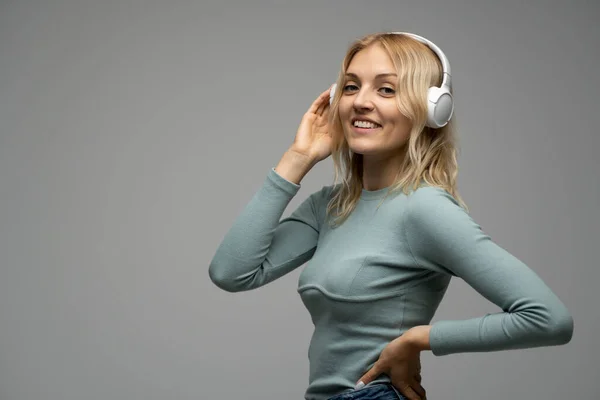 Schöne attraktive junge blonde Frau trägt blaues T-Shirt und Brille mit weißen Kopfhörern, hört Musik und lächelt auf grauem Hintergrund im Studio. Entspannen und genießen. Lebensstil. — Stockfoto