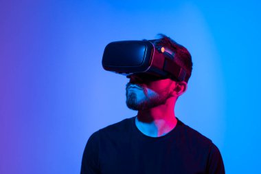 Şaşırmış genç sakallı adam, yüksek teknoloji ürünü VR gözlükler takıyor, 360 derecelik video izliyor ya da metaevrende video oyunu oynuyor.