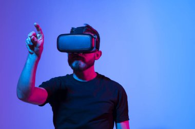 Şaşırmış genç sakallı adam, yüksek teknoloji ürünü VR gözlükler takıyor, 360 derecelik video izliyor ya da metaevrende video oyunu oynuyor.