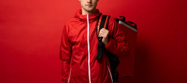 Delivery guy employee man in red uniform workwear work as dealer courier hold red thermal food bag backpack isolated on red color background studio. Service concept