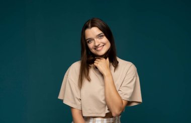 Bej tişörtlü, mutlu, gülümseyen ve pozitif esmer kadın portresi kameraya bakıyor, mavi, turkuaz arka plan stüdyosunda kaygısız gülümsüyor. Duygular