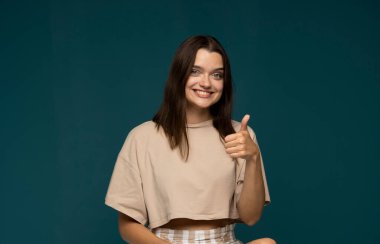 Young brunette woman in a oversized beige t-shirt show thumbs up gesture over cyan wall