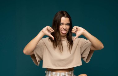 Young brunette woman in a beige t-shirt isolated on cyan background showing thumb down gesture with two hands