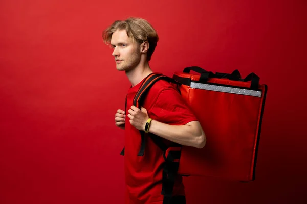 Side view of delivery man in a red t-shirt carry red thermal backpack full of food, groceries to the clients. Food delivery service