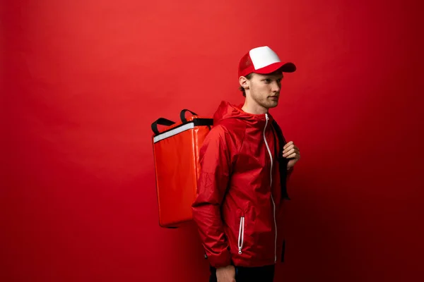 Delivery man in a red jacket and cap carry red thermal backpack full of food, groceries to the clients. Food delivery service