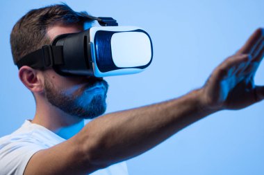 Bearded man in a white t-shirt interacting with the metaverse while wearing virtual reality goggles. Young man using his finger to press a virtual button