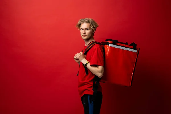 Side view delivery man in a red t-shirt with thermal insulated bag on a shoulder looking in a camera. Food delivery service