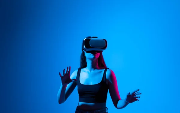 Metaverse technology concept. Confident brunette young woman in virtual reality headset pointing in the air while standing in a studio space. Using VR technologies. Futuristic lifestyle