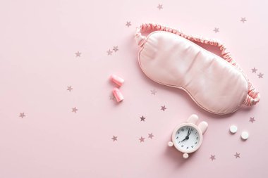 Göz maskesi, çalar saat, kulak tıkaçları, pembe arka planda haplar. Uykusuzluk tedavisi aksesuarları