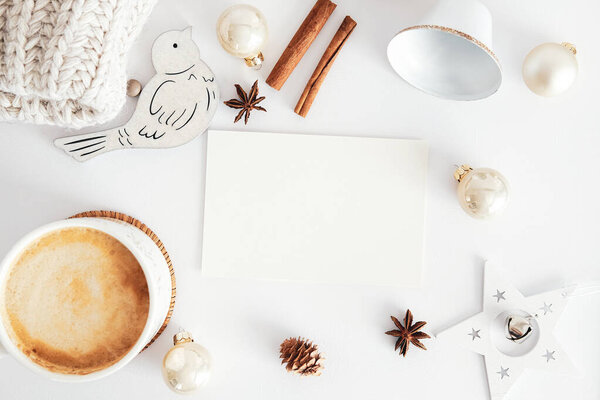 Винтажный макет рождественской открытки. Чистые бумажные карточки, вязаный шарф, коричные палочки, кофейная чашка, бежевые шарики украшения на белом столе. Плоская кладка, вид сверху, копировальное пространство