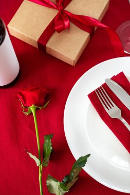 Romantik Sevgililer Günü yemek masası. Çatal bıçak takımı, gül, kırmızı arka planda şarap şişesi..