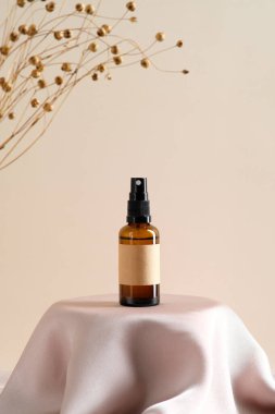 Kahverengi kehribar cam sprey şişesi. Etiket modeli ve bej arka planda kürsüde kuru çiçekler..