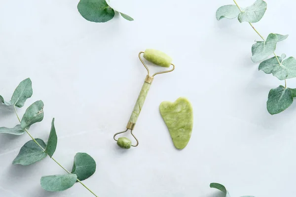 Jade stone facial roller and gua sha on stone table with green eucalyptus leaves. Face skin care concept. Flat lay, top view.
