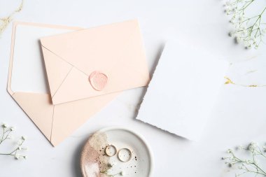 Düğün kırtasiyesinin üst görüntüsü. Düz renkte pembe zarflar, düğün davetiyesi, yüzükler, mermer masada çiçekler..