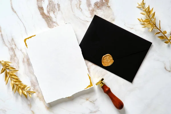 Luxury wedding invitation card mockup with black envelope, gold wax seal stamp, golden leaves on marble gold table. Flat lay, top view, copy space.