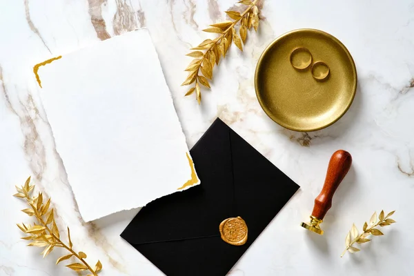 Luxury wedding stationery set on marble gold table. Wedding invitation card mockup, black envelope. gold wax seal stamp, rings, leaves. Top view, flat lay.